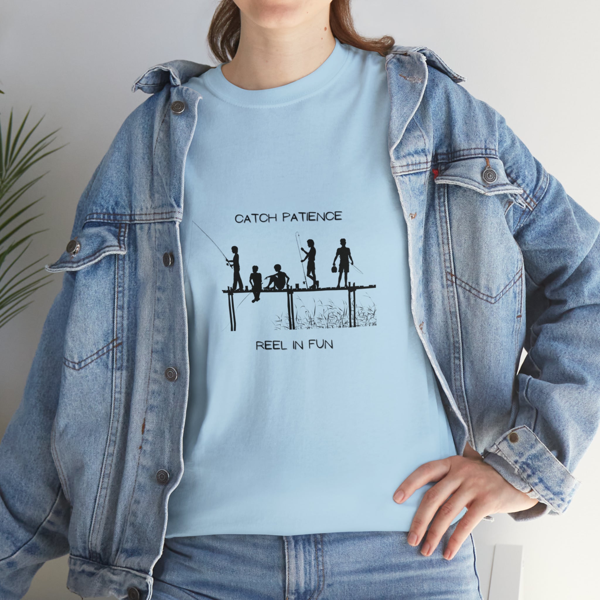 Front view of Adult female wearing light blue T-shirt with "Catch Patience Reel in Fun" inspired graphic of people fishing with jean jacket.