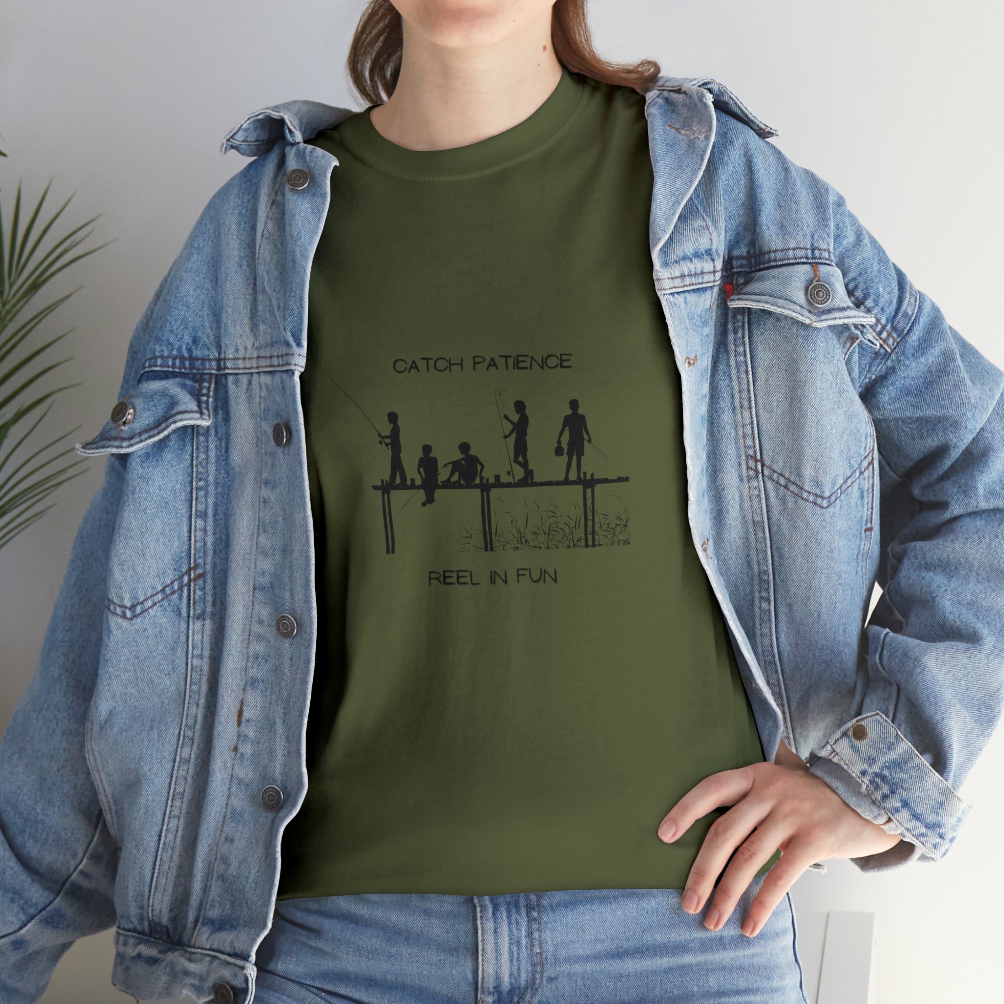Front view of Adult female wearing army green T-shirt with "Catch Patience Reel in Fun" inspired graphic of people fishing with jean jacket.
